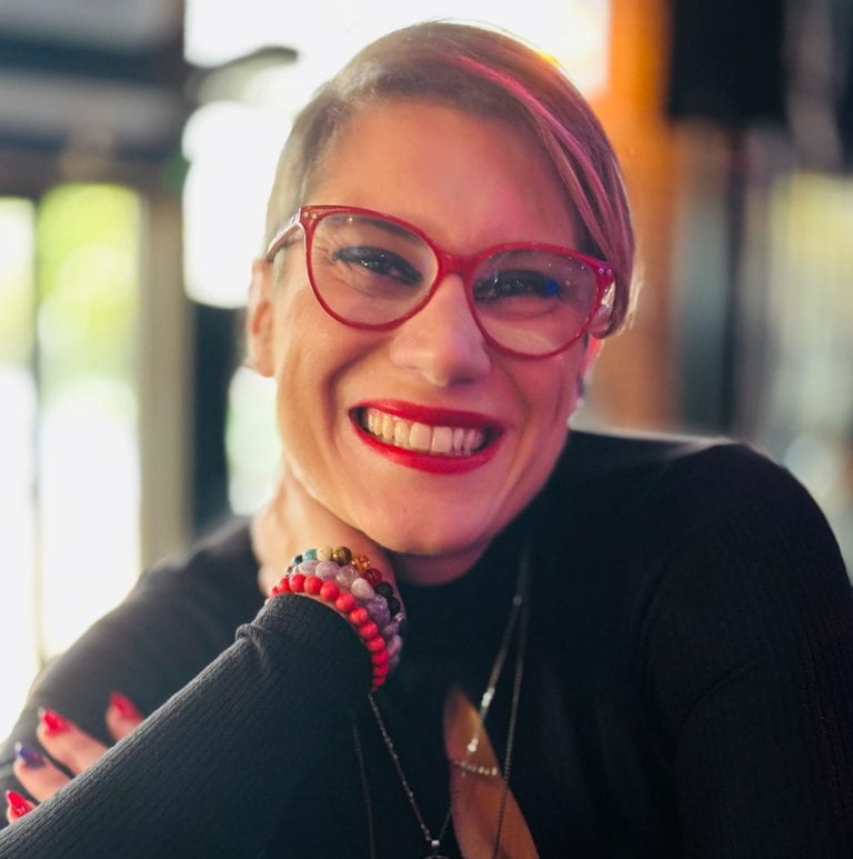 Paola Cavallari, smiles for the camera. She is white, and wearing red glasses and lipstick. Her hair is purple.