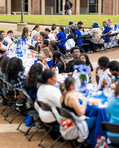 People sit together at a long outdoor table during the Longest Table Event on campus. The table is set with white tablecloths, blue decorations, and floral centerpieces, with academic buildings and walkways visible in the background.