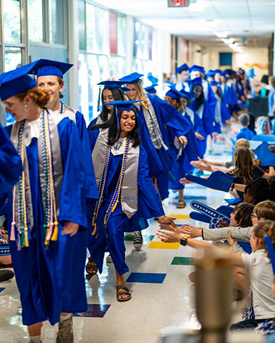 Graduates wearing blue caps and gowns walk down a school hallway, exchanging high-fives with seated students along the sides. The hallway is lined with windows, and colorful floor tiles and wall displays are visible.