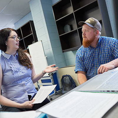 A female and male talking in a classroom near a computer with papers surrounding them