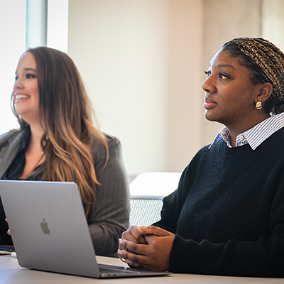 Two females in professional attire and with a laptop anre sitting at a table listening to a lecture