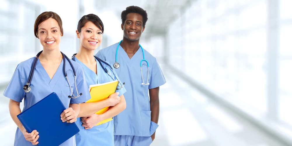 Three healthcare workers dressed in scrubs and wearing stethoscopes around their necks. Two females are holding patient folders